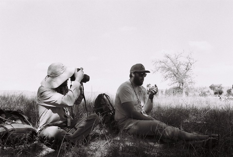 Photograph of two people seated on the ground. The person on the left is taking a photo of the person on the right.