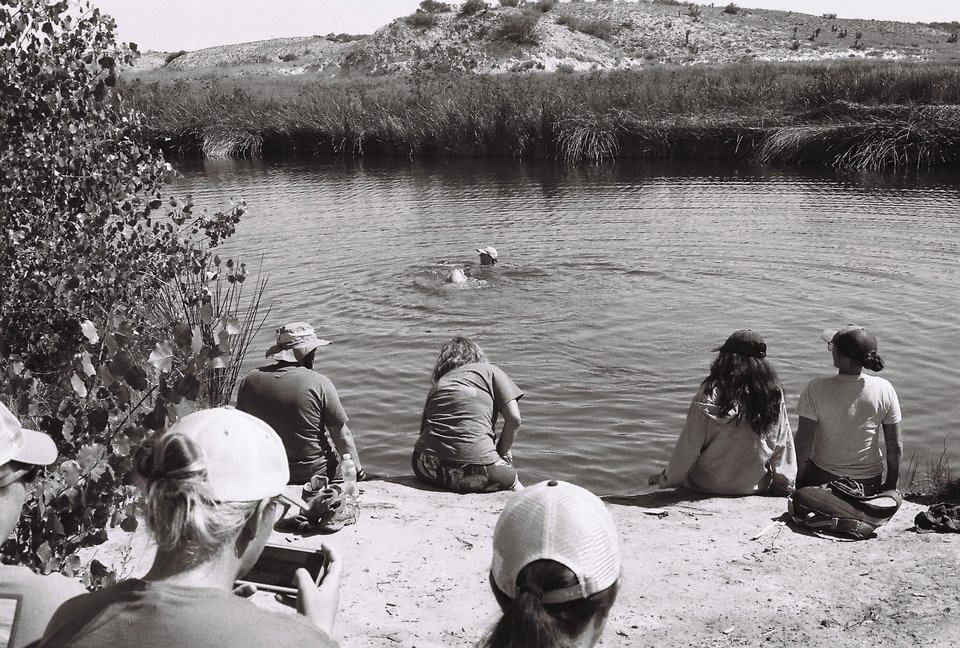 Photograph of several people seated on a riverbank while two people swim in the river.